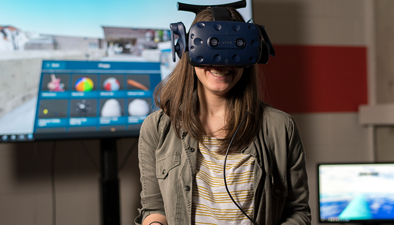 A smiling young woman wearing a virtual reality headset stands in front of a display screen.