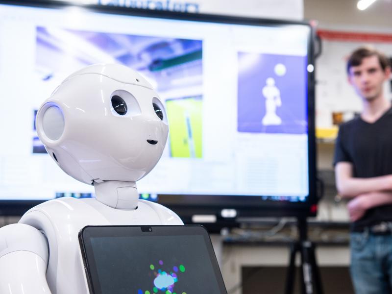 An all-white robot with a very rounded head has a computer screen on its chest.  It is looking very attentively at someone outside the picture frame.  A student stands off to the side watching.